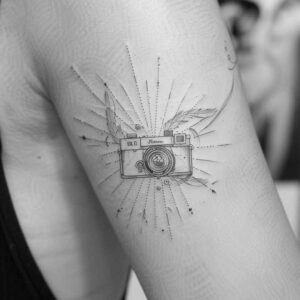 Camera tattoo in black and grey with radiating pattern around the lens; linework project idea, suitable for placement and not specifically a cover-up.
